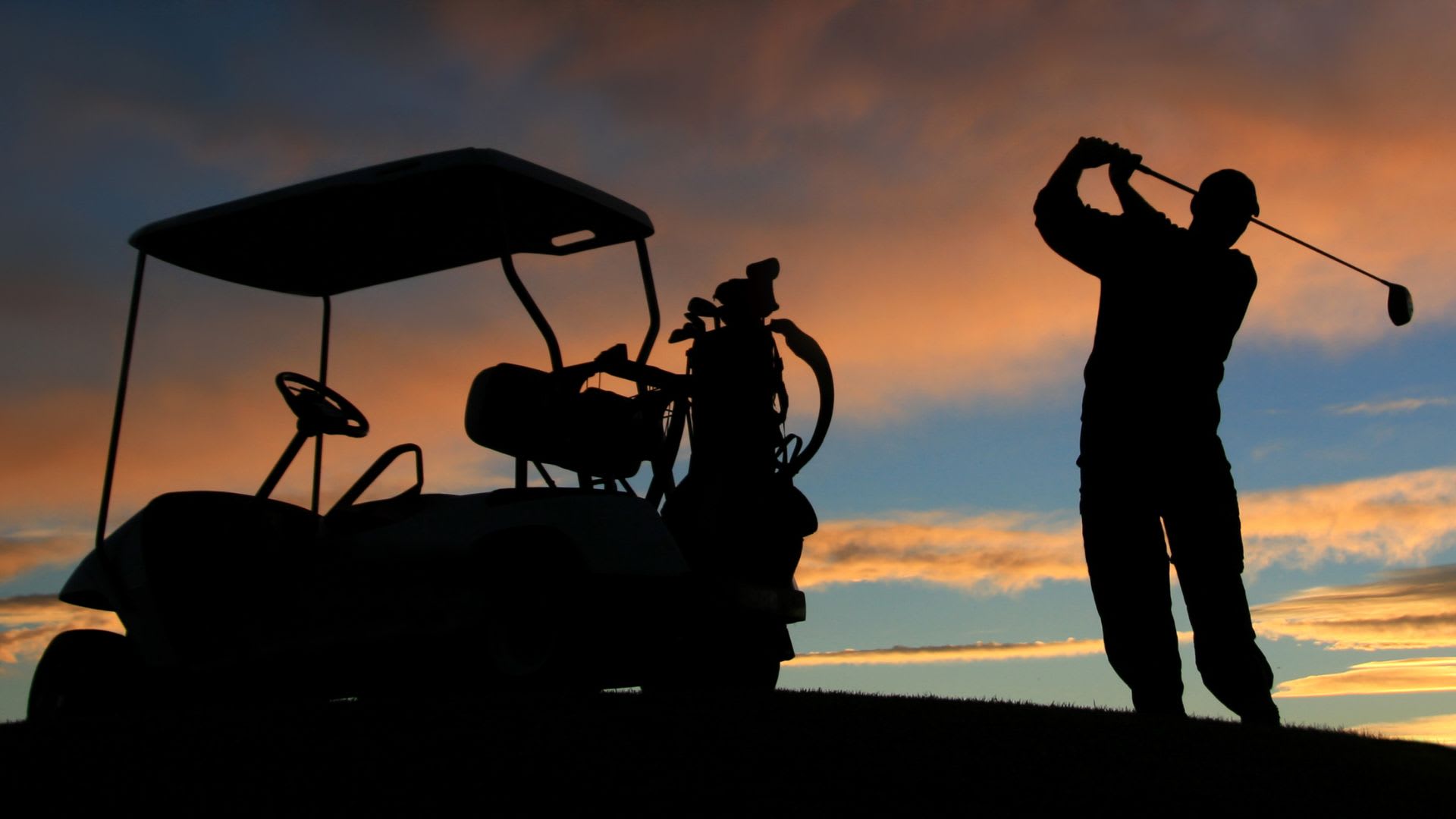 Silhouette of a person swinging a golf club next to a golf cart at sunset with a colorful sky in the background.